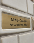 A detail of a donor wall features a gold plaque with the words "Michigan Council for Arts & Cultural Affairs" is surrounded by a white tile border.
