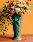 A dramatic bright orange, blush pink, and white floral arrangement with contrasting dark greenery spilling out of the top of a Pewabic Calla Vase in a matte Elm glaze. The vessel is set upon terracotta roof tiles against a bright orange backgrond.