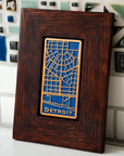 This rectangular Detroit Map Tile has a line drawing of the areal street view of downtown Detroit. The word "Detroit" is written at the bottom of the design. The raised areas of this tile - the word Detroit and the street lines - are scraped which means that it has an orange color while the background is a dark matte blue glaze. The frame is a deep reddish brown oak wood.