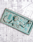 This high relief rectangular tile features a detail of the Spirit of Detroit sculpture found in downtown Detroit. The figure's arms are outstretched with a gilded sphere emanating rays in his left hand and a miniature family in his right. His head is bowed towards the family. The tile has a simple border. This tile features the mottled pale green Patina glaze. It has a similar look to the weathered green finish of copper.