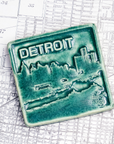 The Detroit Skyline tile features the current downtown skyline with the Detroit River in the foreground. A freighter floats by causing ripples on the otherwise flat surface of the water. The word "Detroit" is written above in the sky. This Detroit Skyline Tile features the matte blueish-green Pewabic Green glaze.