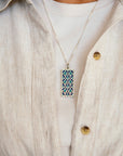The Women's City Club necklace is a rectangular metal pendant with a geometric design that includes teardrop shapes and smoothly curved lines in various shades of blue. The necklace is worn by a person in a light-colored shirt with a linen button-up top.