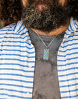 A man wearing a blue and white striped shirt paired with the Women's City Club necklace. The necklace features a rectangular metal pendant with a geometric design that includes teardrop shapes and smoothly curved lines in various shades of blue.