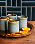 Four ceramic Rocks Cups with a warm beverage, cinnamon sticks, and orange slices on a wooden tray against a dark tiled wall.