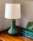 A matte green Elm glazed table lamp with an off-white shade next to a stack of books against a neutral wall.