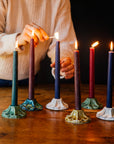 A person in a cream sweater is lighting a row of colorful candles on a wooden surface. The candles are held by decorative, scallopped, floral, ceramic candleholders crafted at Pewabic Pottery in a range of glaze color options from glossy "Honey", matte "Birch" and "Elm", to a semi-reflective "Matte Green Iridescent."