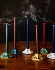 Set of colorful candles that have been recently blown out on a wooden surface against a dark background. The candles are held by decorative, scallopped, floral, ceramic candleholders crafted at Pewabic Pottery in a range of glaze color options from glossy "Honey", matte "Birch" and "Elm", to a semi-reflective "Matte Green Iridescent."
