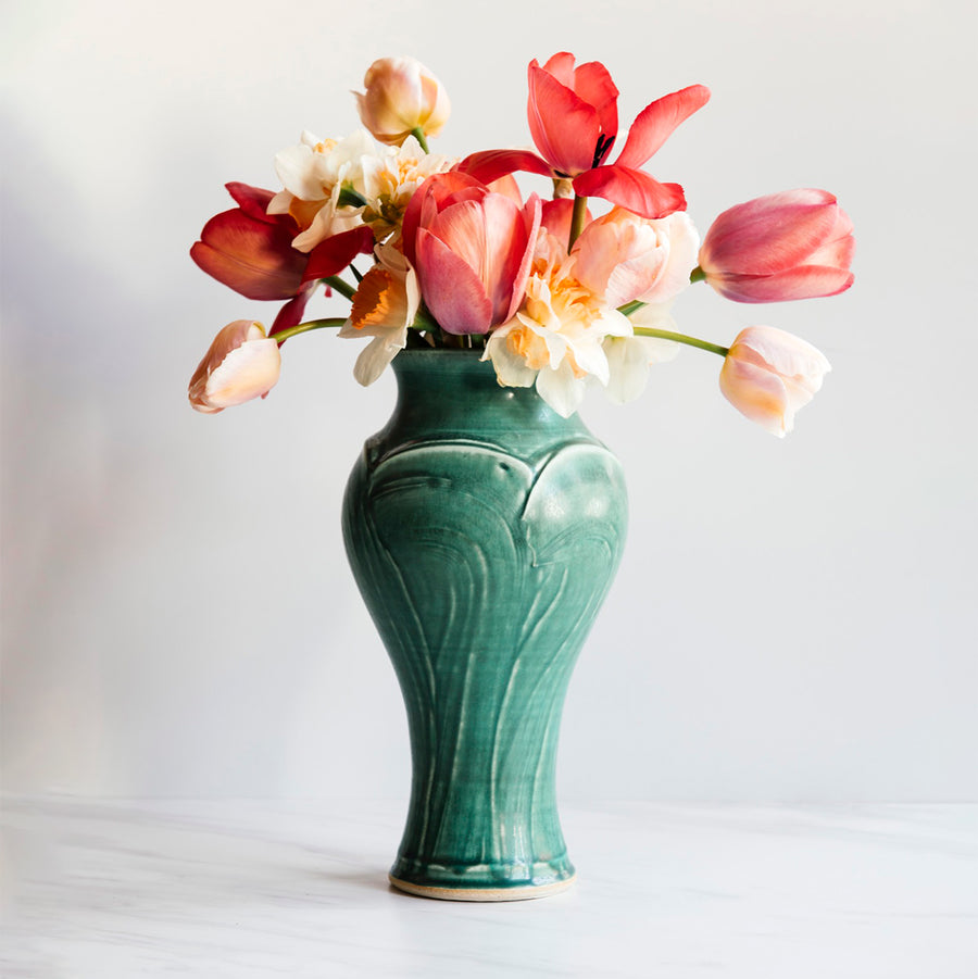 A Pewabic Green-glazed, Large Classic Vase dressed with pink tulips, and neutral daffodils against an ivory backdrop.