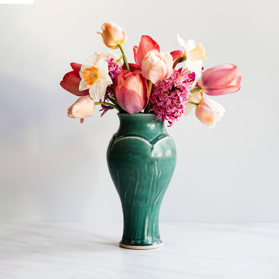 A Pewabic Green-glazed, Medium Classic Vase dressed with pink tulips, and neutral daffodils against an ivory backdrop.