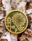 A two-tone glossy, round Sunflower Trivet with golden border and outline set against an emerald green background.