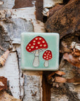 A hand-painted tile depicting a line drawing of a pair of Amanita mushrooms with classic red tops and white spots set on a light sea foam colored background glaze.