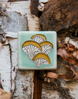 A hand-painted tile depicting a line drawing of Oyster mushrooms with golden yellow tops and white bases set on a light sea foam colored background glaze.