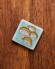 A hand-painted tile depicting a line drawing of Oyster mushrooms with golden yellow tops and white bases set on a light sea foam colored background glaze.