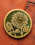 A two-tone glossy, round Sunflower Trivet with golden border and outline set against an emerald green background.