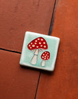 A hand-painted tile depicting a line drawing of a pair of Amanita mushrooms with classic red tops and white spots set on a light sea foam colored background glaze.