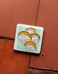 A hand-painted tile depicting a line drawing of Oyster mushrooms with golden yellow tops and white bases set on a light sea foam colored background glaze.