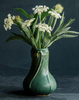 An Elm-glazed ceramic Snowdrop Vase containing a bouquet of white and green flowers, on a dark background.