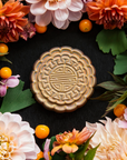 A Mooncake Paperweight in shimmering Blush Iridescent surrounded by vibrant summer florals and gooseberries.