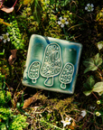 The Morel Tile features three morels - a large one in the center with two smaller mushrroms on either side. The mushrooms have an elongated vertical cap with irregular, sponge-like holes across its surface. The short stems peek out from under the tall cap.