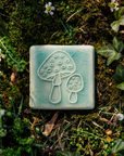 The Amanita Mushroom Tile features a line-drawing of two mushrooms, one small and one large. Each mushroom has polka dot-like circles on its cap and the the larger mushroom is slightly angled, showing off the gills that peek out under its round top.