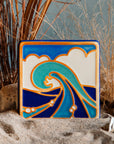 The Wave Tile features a line drawing of a large, curled wave. There are bubbles scattered around its base with a huge puffy cloud behind it that almost covers the bright blue sky. The hand painted tile has multiple blue hues with bright white accents.