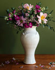 Our Large Classic Vase in a creamy white Birch glaze with brown, iron speckles throughout. The vase is set against a deep, olive-green backdrop with a warm wood foreground. Bright summer flowers decorate the vessel and include a range of purples, soft-pinks, and muted magentas.