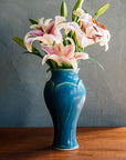 Our bright, medium, blue Peacock glaze on the Large Classic Vase form. This vessel is overflowing with pink lilies. The background is a textured, lime-washed dark grey and the foreground a warm wood finish.