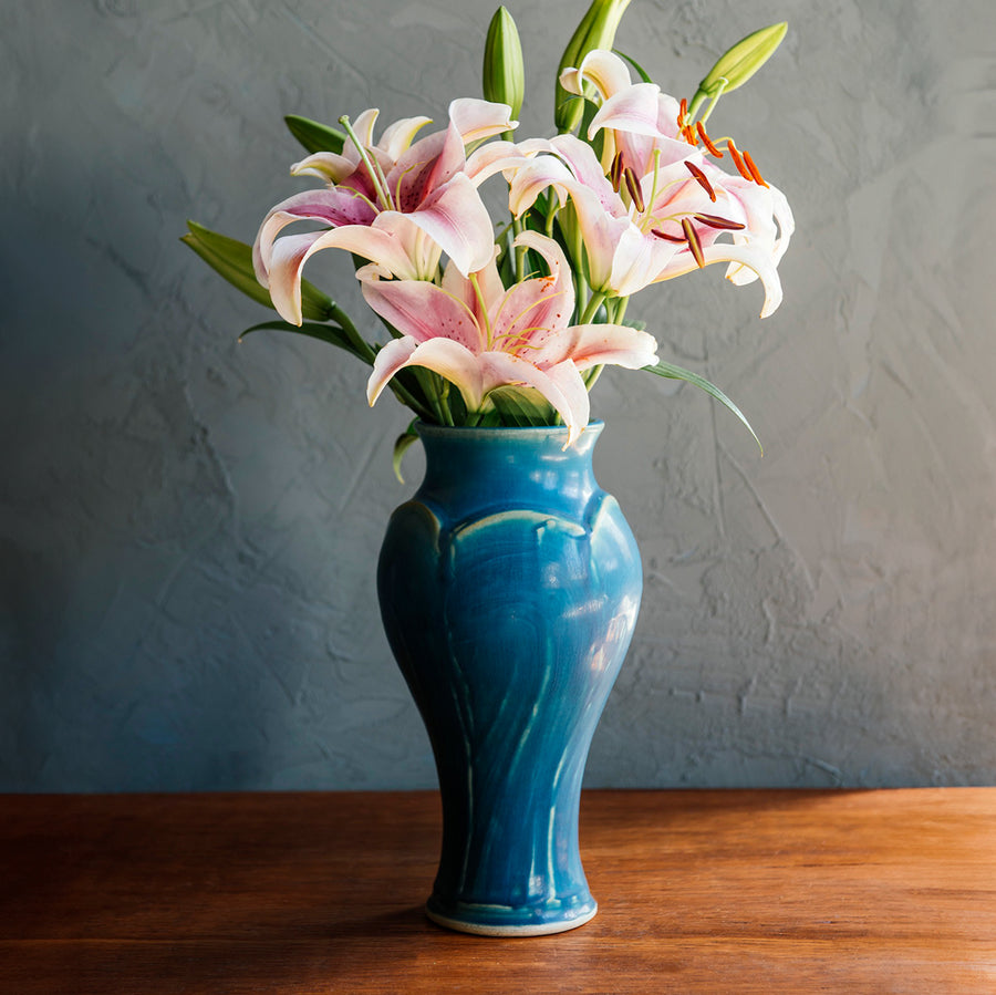 Our bright, medium, blue Peacock glaze on the Large Classic Vase form. This vessel is overflowing with pink lilies. The background is a textured, lime-washed dark grey and the foreground a warm wood finish.