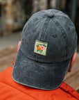 Person wearing a dark gray cap with a floral patch, sitting outdoors.