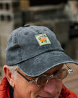 Man wearing a dark gray cap with a floral patch and sunglasses, outdoors.