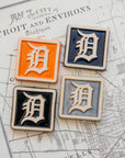 Four Old English 'D' logo tiles in a range of colorful glaze options on a map of Detroit, Michigan.