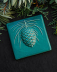 The 6x6 Pinecone tile features a large pinecone in the center of the tile with pine needles encircling it. This piece shown in Pewabic blue- a turquoise blue color.