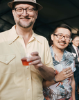 Two men smile with cocktails in hand during the Pewabic Preview Party Fundraiser. The man on the left has a butter yellow shirt and hat, while the man on the right sports a palm frond patterned button up shirt