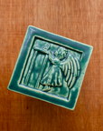 The Little Angel Girl Tile features a a toddler in a long nightgown with large feathered wings. She is on tip toes reaching for a lit candle on a high table. The candle is pulled towards her and the flame is wavering from the breeze created as she tries to blow out the flame. This tile features the matte blueish-green Pewabic Green glaze.