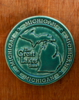 This round Michigan Trivet Tile features the outline of the state's two peninsulas including some of the larger northern islands of Michigan. To the left of the design reads "The Great Lakes State" and surrounding it, in a border, reads the word Michigan four times.