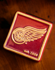 The bright red square Red Wings Tile features the Red Wings logo raised in its center and the iridescent quality gives the design a golden hue. There is a simple line border around the tile that is also lighter. In the bottom right corner are the words "est. 1926".