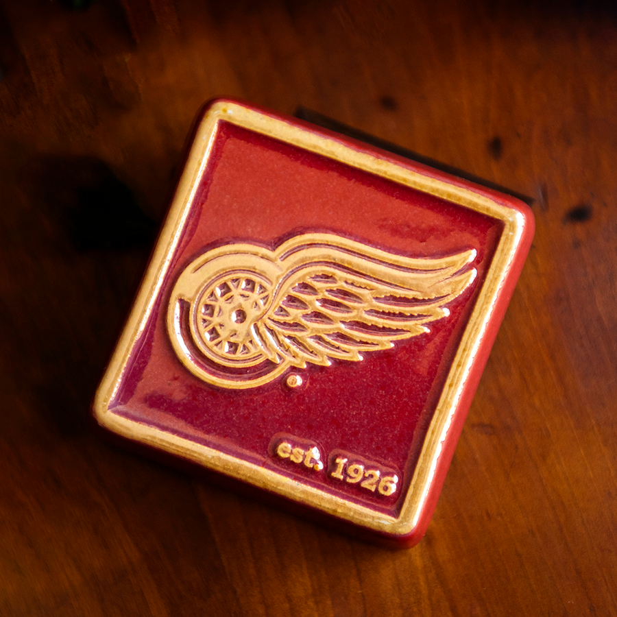 The bright red square Red Wings Tile features the Red Wings logo raised in its center and the iridescent quality gives the design a golden hue. There is a simple line border around the tile that is also lighter. In the bottom right corner are the words "est. 1926".