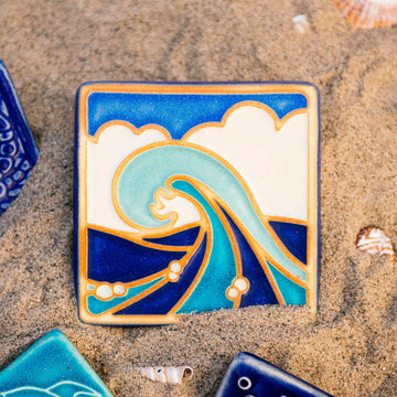 The Wave Tile features a line drawing of a large, curled wave. There are bubbles scattered around its base with a huge puffy cloud behind it that almost covers the bright blue sky. The hand painted tile has multiple blue hues with bright white accents.