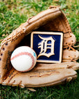 A leather baseball mitt is nestled in bright, green grass. Inside the mitt lays a baseball and our 4x4" Tigers Old English "D" Tile in a Navy Blue and light "Natural" clay border. The Old English "D" logo is also outfitted in the light clay body, allowing it to stand out against its bright backdrop.