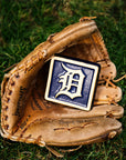 A leather baseball mitt is nestled in bright, green grass. Inside the mitt lays a our 4x4" Tigers Old English "D" Tile in a Navy Blue and light "Natural" clay border. The Old English "D" logo is also outfitted in the light clay body, allowing it to stand out against its bright backdrop.