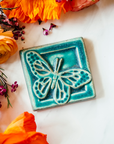 The ceramic Butterfly tile's design sits diagonally on the 4x4 square tile. Its wings are outstretched and it is floating above a depressed border that surrounds the tile. This gives the illusion that the butterfly is flying out toward you. This tile features the matte turquoise Pewabic Blue glaze.