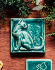 A tile sits on a wooden table surrounded by greenery. The matte green, Pewabic Green embossed tile shows a sleek cat sitting up straight with its head turned backward towards its flicking tail.