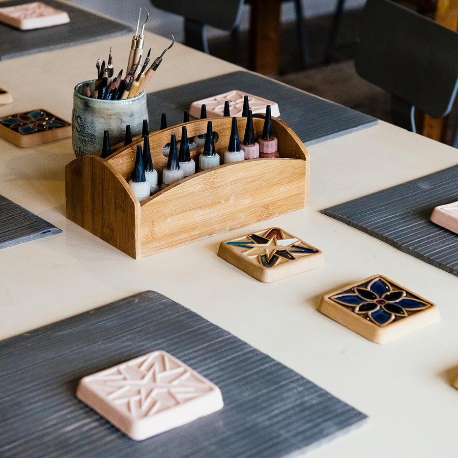 A table is set up with all the supplies needed to glaze an unfinished tile; a caddy full of small bottles of glaze, a cup full of small paint brushes, unfinished  clay tiles and examples of completes pieces. 