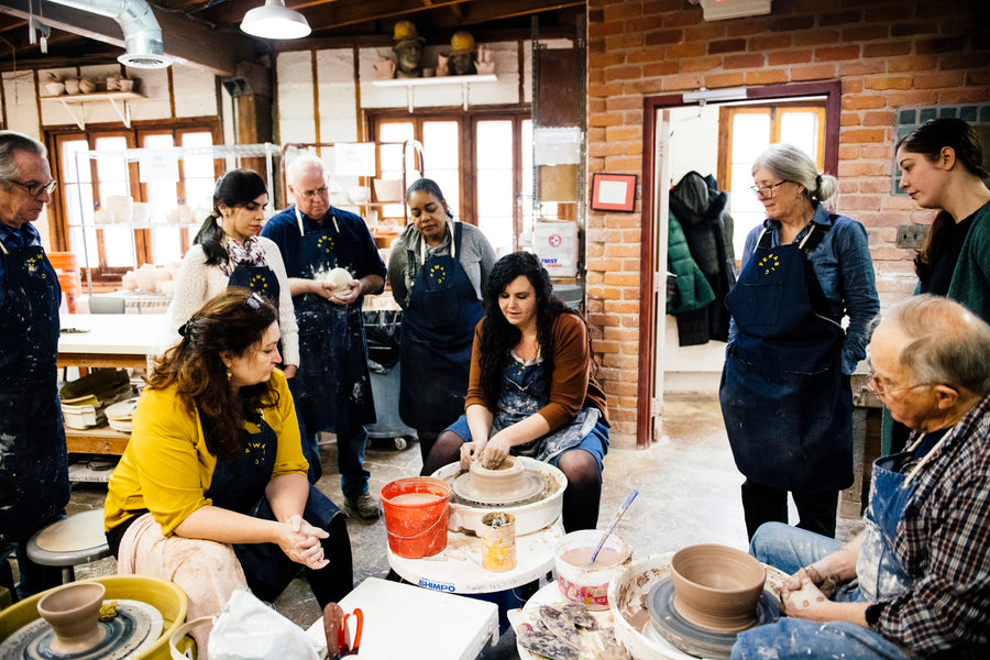 A Pewabic educator sits at a pottery wheel in our education studio. She molds a cup out of wet clay as her students watch to learn her technique.