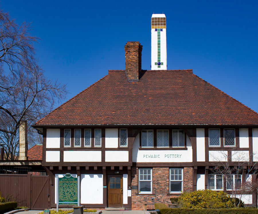 The historic pottery building features white stucco walls are bordered by brown wooden beams. The terracotta tile roof is punctuated by a tall white chimney that features colorful Pewabic tiles.