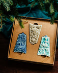 Three ceramic ornaments are sitting in a gift box and placed under a Christmas tree.