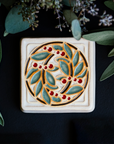 The DIA Leaf and Berries tile features two intertwined branches with round berries that create a circle at the center of the tile. The thick lined border makes the tile into a square. This tile features Style D with red berries, pale green leaves, brown stems and a white background.