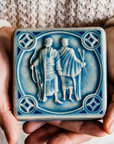 The glossy pale blue Celestite glaze pools across the low-relief sculpted twin forms of this Gemini tile. Two warm hands hold the tile in natural light to show off the glossy surface.