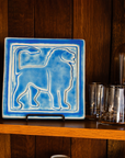 This Aztec Lion tile features the matte bright blue Sky glaze. The lion faces to the right and rests on the wooden bookshelf of a collector.