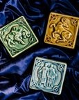 The Gemini tile lies with the other two Pewabic Zodiac tiles on rich, velvety fabric the color of the night sky. The Taurus tile features a sturdy bull with its head bowed and horns lowered. The Aries tile has an alert ram with its legs positioned as if it is prancing. The ram's head is turned backwards over its shoulder.